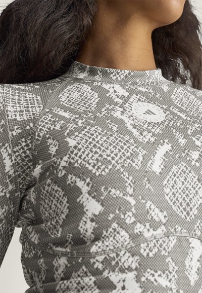Primer plano de una persona que lleva una camisa gris y blanca con un estampado, con un logo redondo cerca del cuello y cabello oscuro y ondulado.
