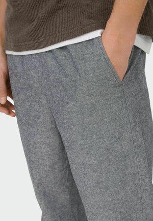 Moški, oblečen v sive teksturirane hlače z roko v žepu in rjav pulover iz pletenine čez belo srajco, prikazan od pasu do sredine stegna.