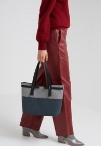 Persona che indossa pantaloni di pelle rossi e un maglione bordeaux, mentre porta una borsa a tracolla in pelle grigia e blu navy con manici neri.