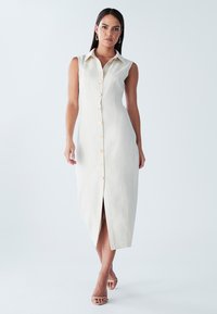 Mouwloze linnen jurk in lichtbeige met een knoopsluiting aan de voorkant en een hoge split aan de zijkant. Voorzien van een kraag en een subtiele textuur.