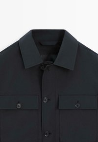 Czarna kurtka z kołnierzem, zapinana na guziki z przodu, posiadająca dwie kieszenie na piersi zamykane na guziki, przedstawiona na białym tle.