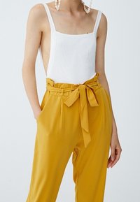 Vit ribbad bodysuit med fyrkantig halsringning som kombineras med högmidjade gula byxor med knytbälte och sidfickor.