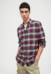 Piros és tengerészkék kockás ing hosszú ujjal, a mandzsettáknál felgyűrve, sima textúrájú anyagból készült. Khaki nadrággal párosítva.