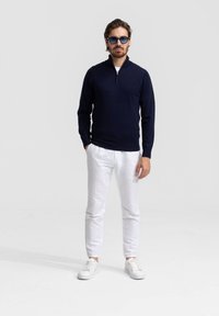 Marine ritsvest, witte broeken en witte sneakers. Het vest heeft een gladde textuur; de broeken zijn van lichtgewicht stof; de zonnebril heeft een blauwe tint.