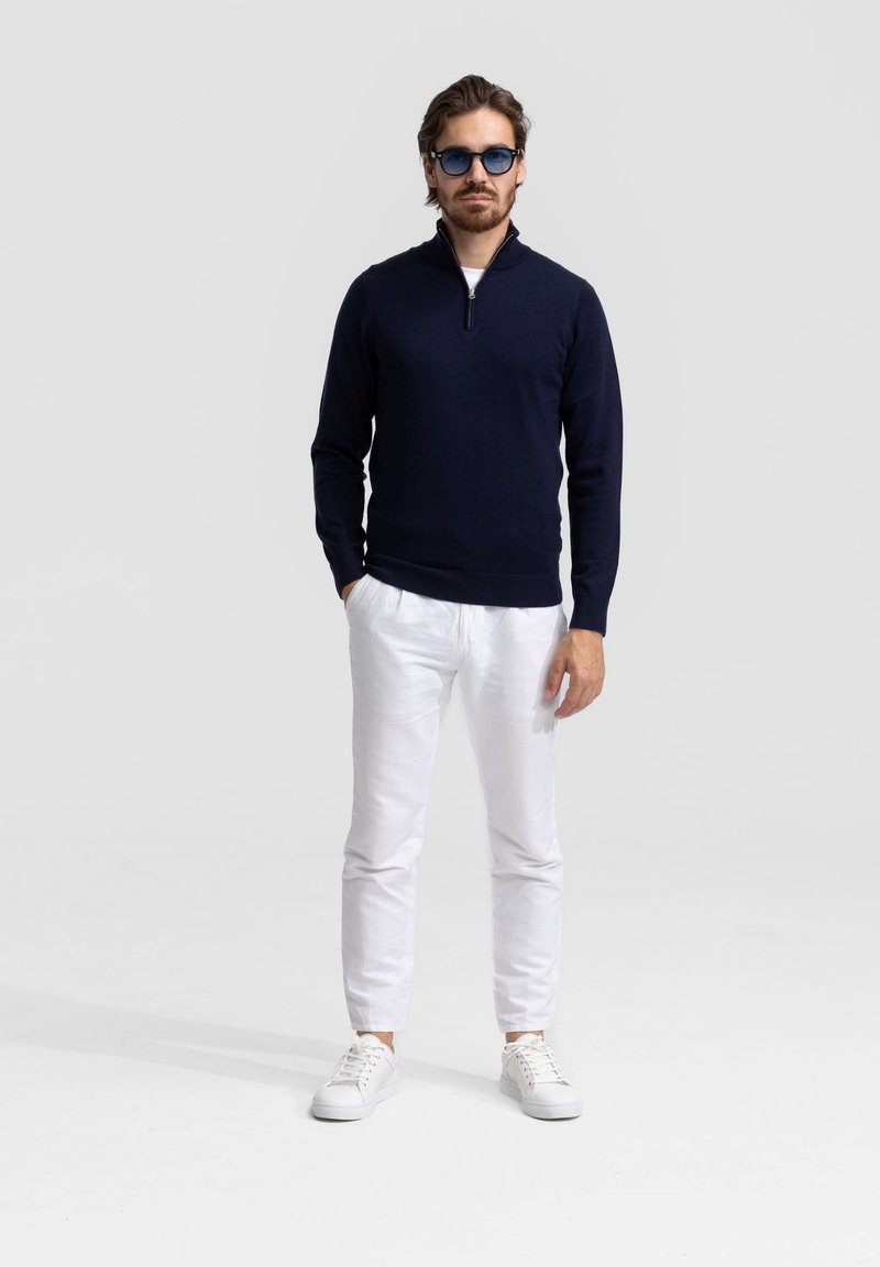 Marine ritsvest, witte broeken en witte sneakers. Het vest heeft een gladde textuur; de broeken zijn van lichtgewicht stof; de zonnebril heeft een blauwe tint.