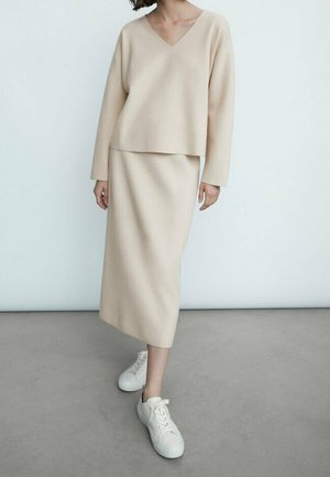 Beige neulepaita V-pääntiellä ja pitkillä hihoilla, yhdistetty samanväriseen polvipituiseen hameeseen, valkoisten tennareiden kanssa harmaalla lattialla.