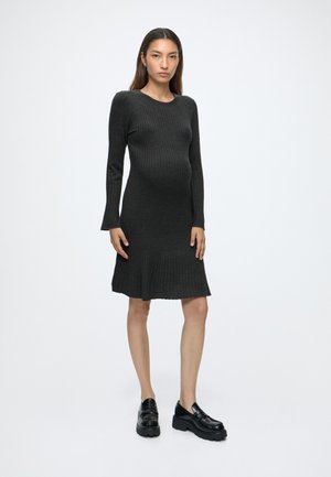 Femme aux longs cheveux portant une robe noire en maille côtelée et des mocassins noirs épais, debout devant un fond blanc uni.