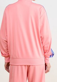 Rosa träningsoveralljacka med hög krage, ribbade ärmslut och midja. Har blå ränder längs ärmarna, slät textur.