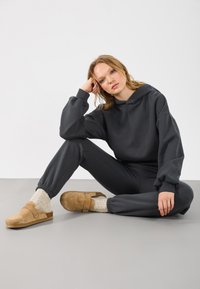 Sudadera gris oscuro con mangas anchas y joggers a juego. Zuecos de ante beige usados con calcetines de punto color crema. Textura de tela suave y lisa.