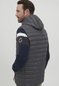 Siv voden jopič s kapuco, ki ima temno modre pletene rokave in bel strižen akcent. Vključuje okroglo logo na rami.