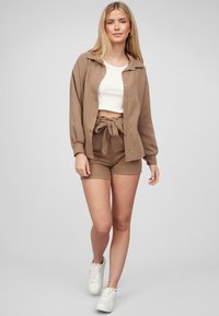 Camicia button-up marrone con colletto, top bianco corto e pantaloni corti marroni con vita regolabile. Indossata con sneakers bianche. Texture liscia.