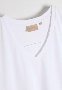 Biała koszulka polo z dekoltem w serek, wykonana z miękkiego, gładkiego materiału. Posiada minimalistyczną, beżową etykietę z rozmiarem "S" oraz proste szwy wzdłuż dekoltu.