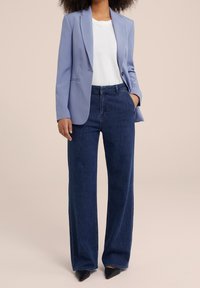 Jasnoniebieska marynarka, biała koszulka i ciemnoniebieskie szerokie jeansy. Buty są czarne z szpiczastymi noskami. Gładki materiał i przemyślany krój.