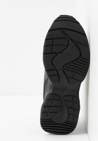 Suola di scarpa sportiva nera che mostra una gomma testurizzata, un motivo ondulato e scanalature profonde per la trazione. Design minimalista senza loghi visibili.