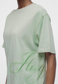 Άτομο που φοράει ανοιχτό πράσινο φαρδύ T-shirt με μεγάλο ανάγλυφο κείμενο με πλάγια γραφή στην πλευρά που γράφει "Aug".