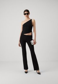 Top nero a spalla unica abbinato a pantaloni neri aderenti. Il top presenta un design arricciato e si annoda di lato. Gli accessori includono occhiali da sole neri e una clutch trasparente.