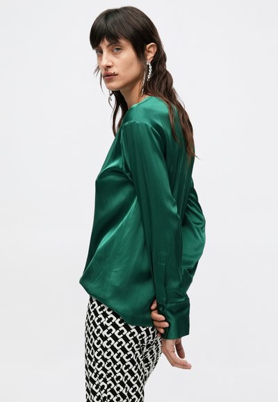 Πράσινο σατέν μπλουζάκι με μακριά μανίκια και άνετη εφαρμογή, συνδυασμένο με εμπριμέ μαύρο και λευκό παντελόνι. Ο μεταξένιος υφασμάτινος χαρακτήρας και η λεπτή λάμψη είναι εμφανείς.