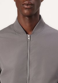 Šedá bomber bunda se zipem vpředu, žebrovaným límcem a hladkým povrchem látky. Bez vzorů; obsahuje minimalistické designové prvky.