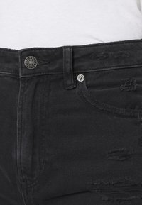 Jean en denim noir avec des détails déchirés, cinq poches, matériel en argent et fermeture à bouton sur une taille texturée.