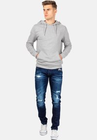 Felpa grigia con cappuccio e tasche a marsupio, abbinata a jeans blu strappati e sneakers bianche. Il tessuto è morbido, con una vestibilità casual.