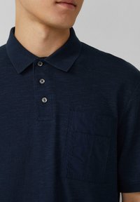Granatowa koszulka polo z teksturowanej tkaniny, kołnierzykiem i trzema guzikami. Posiada jednolitą granatową kieszonkę na lewej piersi.