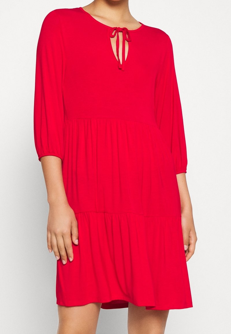 Femme portant une robe rouge vif longueur genou avec des manches trois-quarts et un détail noué au niveau de l'encolure sur un fond blanc.