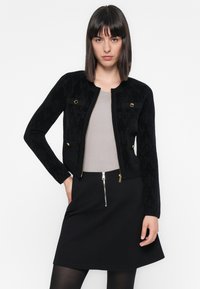 Femeie cu păr drept și închis la culoare, purtând o jachetă neagră cu fermoar și nasturi aurii, un top gri cu dungi, o fustă neagră cu fermoar argintiu și ciorapi negri.