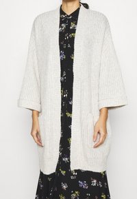 Cardigan lavorato a maglia grigio chiaro con apertura frontale, spalle cadute, texture a coste e due tasche frontali. Indossato sopra un vestito nero floreale.