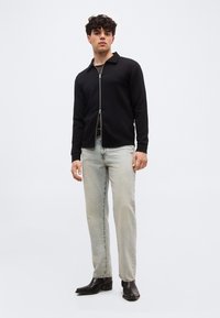 Νέος άνδρας που φοράει μαύρο μπουφάν με φερμουάρ, ανοιχτό μπλε τζιν και μαύρα δερμάτινα μποτάκια, στέκεται μπροστά σε ένα απλό φωτεινό φόντο.