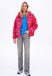 Puszysta kurtka w różowo-pomarańczowy wzór z kapturem, noszona na niebieskim swetrze, w połączeniu z szarymi dżinsami i białymi trampkami.