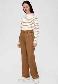 Bruine high-waisted broek met dubbele knoopsluiting en plooien, gecombineerd met een crèmekleurig en bruine gestreepte lange mouw shirt. Sneakers maken de look compleet.