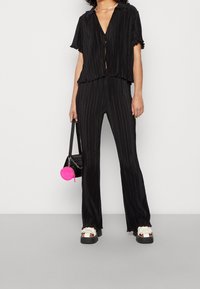 Zwart geplooid twee-delig outfit met korte mouwen, een cropped top en uitlopende broek, gecombineerd met chunkywitte sandalen en een roze ronde tas.