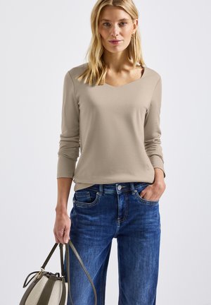 Beige top met lange mouwen en een V-hals, gecombineerd met donkerblauwe jeans. Het model houdt een handtas in twee tonen met groene accenten.