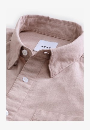 Vaaleanpunainen puuvillapaita, jossa on rakennettu kaulus, valkoiset napit ja etiketti, jossa lukee "NEXT." Paidan pinnassa on teksturoitu kangas.