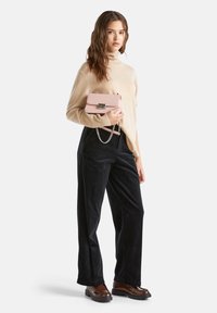 Femme portant un pull col roulé beige, un pantalon large noir, des chaussures à plateforme marron, tenant un petit sac à main rose avec une bandoulière en chaîne.