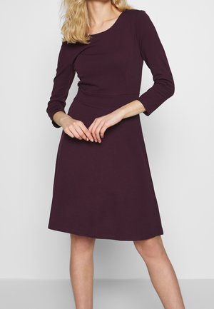 Frau trägt ein knielanges, langärmliges, dunkelviolettes, figurbetontes Kleid und steht vor einem schlichten hellgrauen Hintergrund.