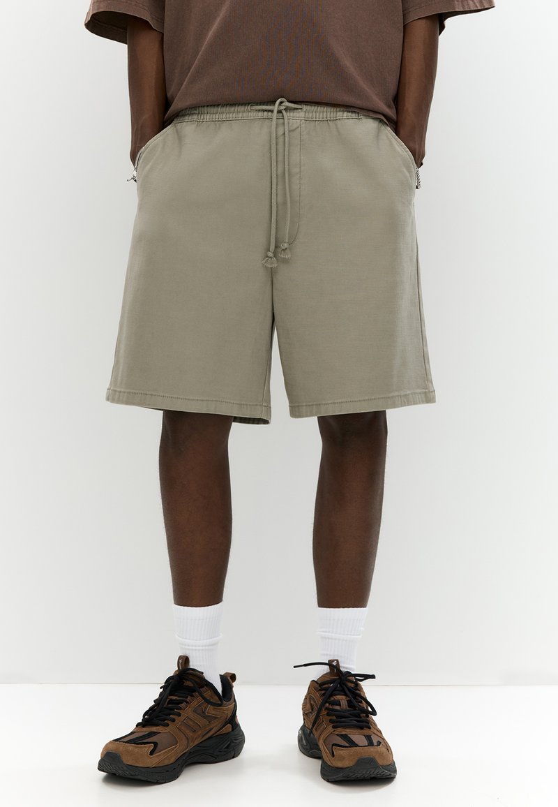 Pantaloni corti in cotone grigio chiaro con vita elasticizzata e cordoncino, dotati di tasche laterali e un fit rilassato. Abbinati a sneakers marroni.