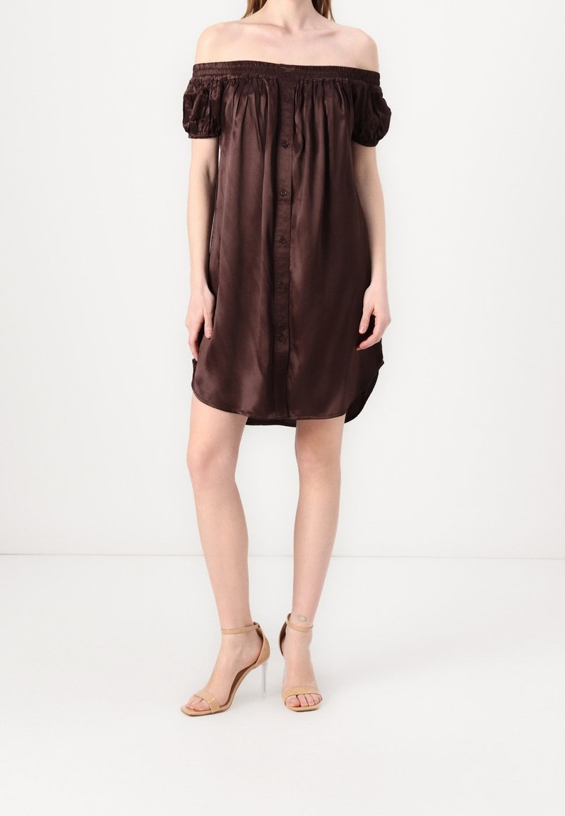 Femme portant une courte robe marron foncé épaules dénudées avec des détails à boutons et des sandales à talons nude, debout devant un fond uni.