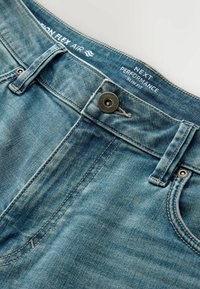 Jean slim fit en denim bleu avec fermeture par bouton en métal et passants de ceinture, avec les marques "Next Performance" et "Motion Flex Air" à l'intérieur de la ceinture.
