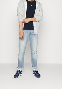 Light gray zip-up hoodie over a navy t-shirt, paired with light blue jeans and navy sneakers, featuring white accents and rolled cuffs.