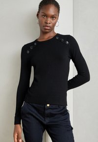 Černý pletený svetr s kulatým výstřihem a dekorativními knoflíky na ramenou. Materiál vypadá hladce a má přiléhavý střih.