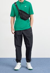 Man med grön Nike-polo, svarta cargobyxor, vita sneakers med gröna detaljer och en svart crossbody-väska på blå matta.