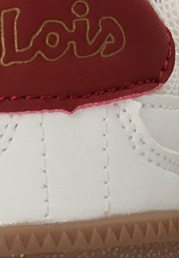 Zapatilla blanca con acentos rojos, que cuenta con una parte superior texturizada, logo dorado y costuras detalladas. La suela es marrón con un patrón de zigzag.