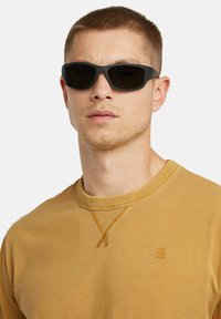 Gli occhiali da sole presentano montature nere con lenti scure. Il modello indossa una felpa color senape con scollo tondo e cuciture testurizzate.