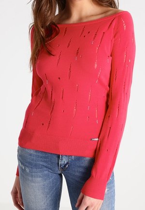 Femme portant un pull en tricot rouge usé à manches longues et un jean bleu déchiré, cadrée sous le menton.