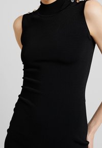 Top senza maniche nero con collo alto e trama a coste. Presenta bottoni decorativi dorati sulla spalla per un tocco di dettaglio e accentuazione.