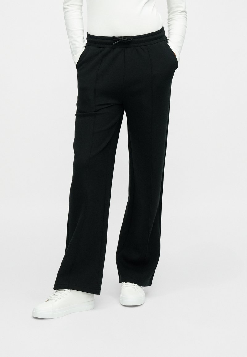 Μαύρο, άνετο παντελόνι φόρμας με κορδόνι στη μέση και πλαϊνές τσέπες. Ομαλή υφή, σχεδιασμός με ίσια μπατζάκια, συνδυασμένο με λευκά αθλητικά παπούτσια.