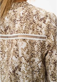 Blus med långa ärmar i en beige och brun ormskinnsdesign, med en vit spetsdetalj längs den övre ryggen. Mjuk tygtextur.