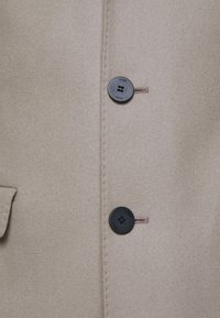 Cappotto di lana beige con una texture liscia, due bottoni neri con logo e dettaglio della tasca laterale. Design sartoriale con bordi cuciti.