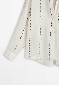 Camicia bianca in lino con motivi ricamati neri verticali, maniche lunghe, scollatura a V e vestibilità rilassata con spacco laterale.